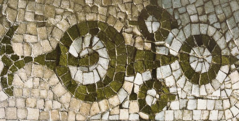 Nahaufnahme eines historischen Mosaiks aus kleinen Steintesserae in Beige-, Weiß-, Grau- und Olivgrüntönen. Das ornamentale Muster wird von zwei spiralförmigen Motiven und geschwungenen Linien geprägt, die sich über die gesamte Bildfläche ziehen. Die unregelmäßigen Fugen und die leicht verwitterte Oberfläche betonen die handwerkliche Struktur und das Alter des Mosaiks.