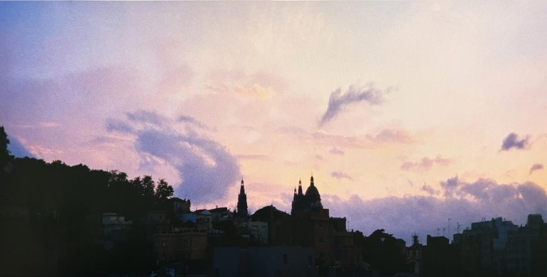 Blick auf die Silhouette Barcelonas bei Sonnenuntergang. Vor einem Himmel in Rosa-, Violett- und Goldtönen zeichnen sich Kirchtürme und Dächer scherenschnittartig ab, während Wolken die stimmungsvolle Abendatmosphäre prägen.