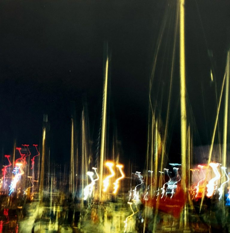 Nachtaufnahme eines Hafens mit Segelmasten, die durch Bewegungsunschärfe als helle, vertikale Lichtspuren erscheinen. Bunte Lichter von Booten und der Uferpromenade zeichnen geschwungene Linien vor dunklem Himmel und erzeugen eine dynamische, malerische Atmosphäre.