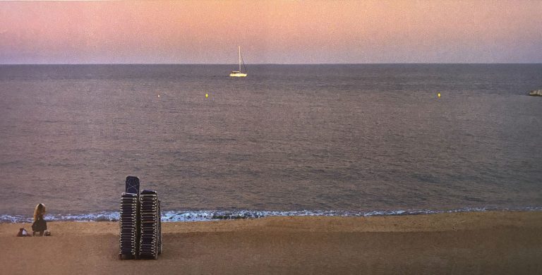 Blick auf einen ruhigen Strand in der Abenddämmerung. Im Vordergrund stehen gestapelte Liegestühle auf dem Sand, links sitzt eine einzelne Person am Ufer. Das dunkle Meer erstreckt sich bis zum Horizont, wo ein Segelboot vor einem Himmel in zarten Rosa-, Orange- und Violetttönen ankert. Die ruhige Komposition vermittelt eine stille, entspannte Atmosphäre kurz nach Sonnenuntergang.