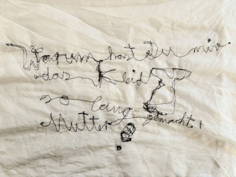 Handgestickter Schriftzug aus schwarzem Faden auf weißem zerknittertem Stoff mit freier Linienführung und kleinen Zeichenelementen: Warum hast Du mir das Kleid so lang gemacht, Mutter?