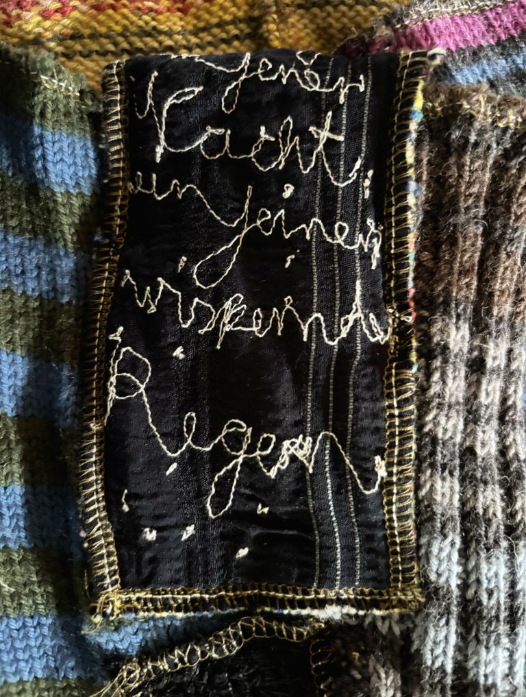 Detail eines textilen Objekts mit handgesticktem Schriftzug auf schwarzem Stoff, eingefasst von bunten Strickflächen.